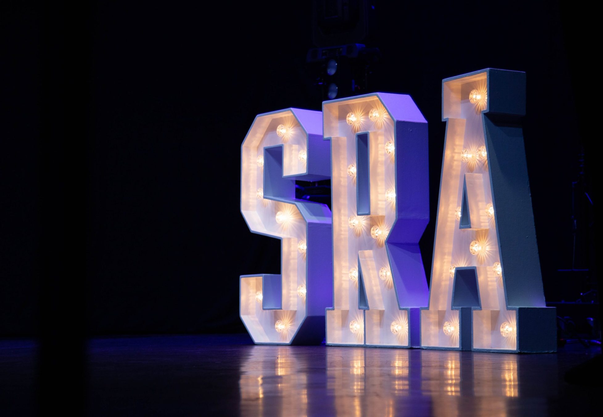 Large illuminated letters spelling S R A, on a stage